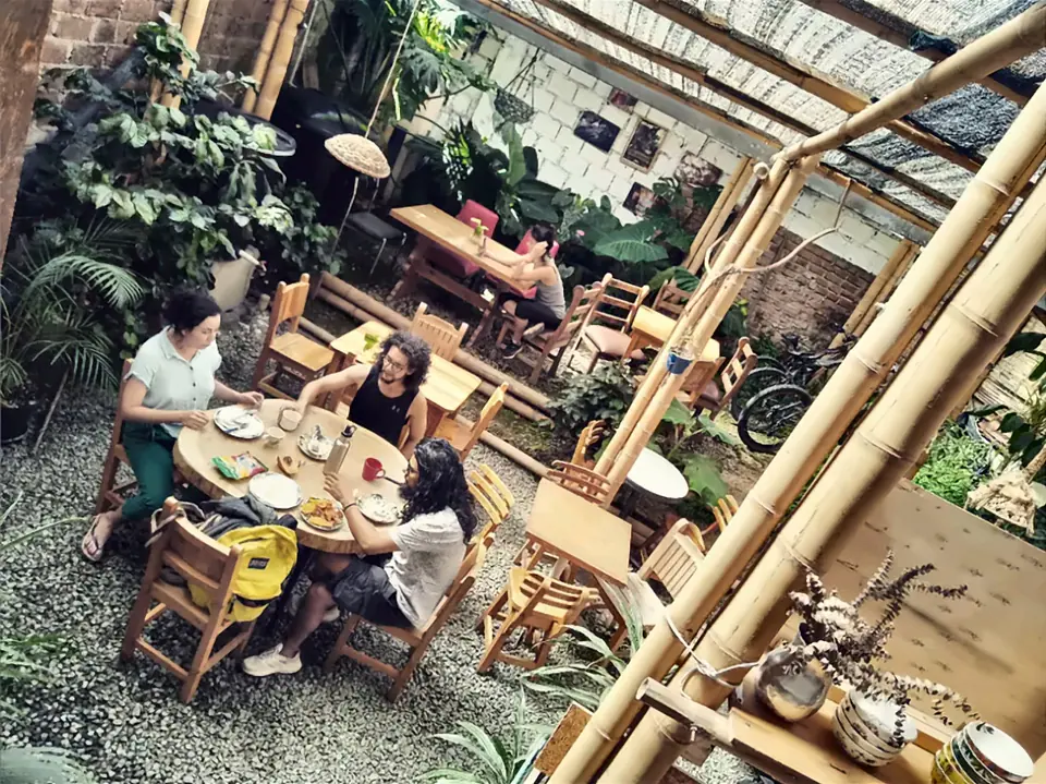 Vista desde arriba del patio de Bidea: viajeros desayunando en una mesa redonda de madera entre plantas y estructuras de guadua