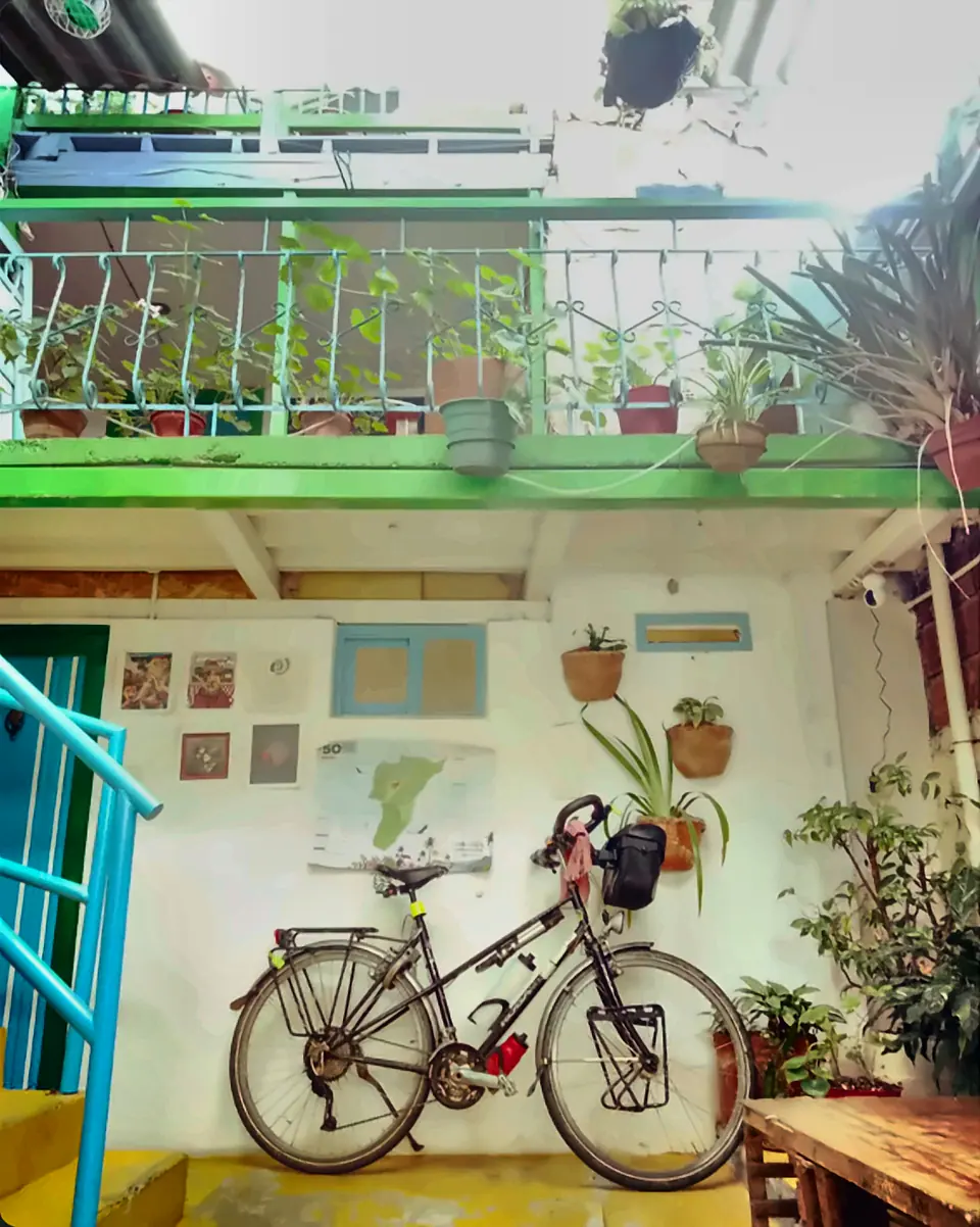 Bicicleta de viaje con alforjas apoyada en una pared del patio interior, bajo un balcón verde con plantas y un mapa colgado