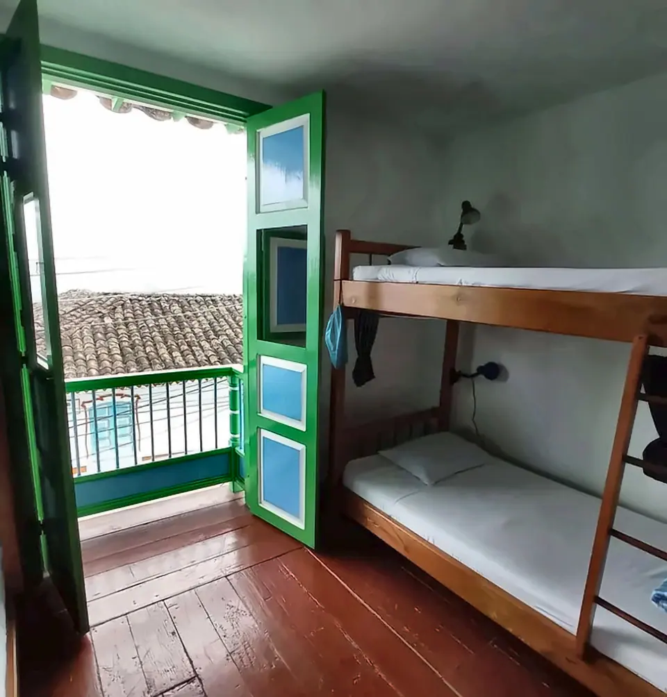 Litera de madera junto a una puerta-balcón verde de doble hoja abierta a un balcón con baranda y tejados de barro