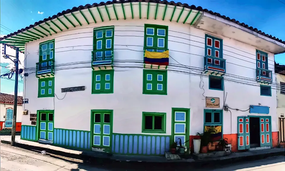 Fachada de Bidea: casa blanca de esquina con ventanas y balcones verdes, vidrios turquesa y rojos, alero de rayas verdes y blancas y una bandera de Colombia en el segundo piso