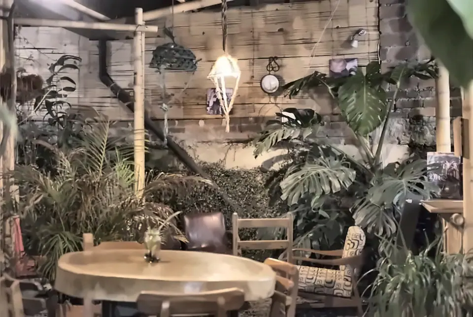 Patio de noche con una lámpara colgante encendida, plantas tropicales, postes de guadua y una mesa redonda de madera